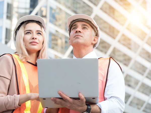 A man and a woman holding a laptop. They are wearing hardhats and reflective vests