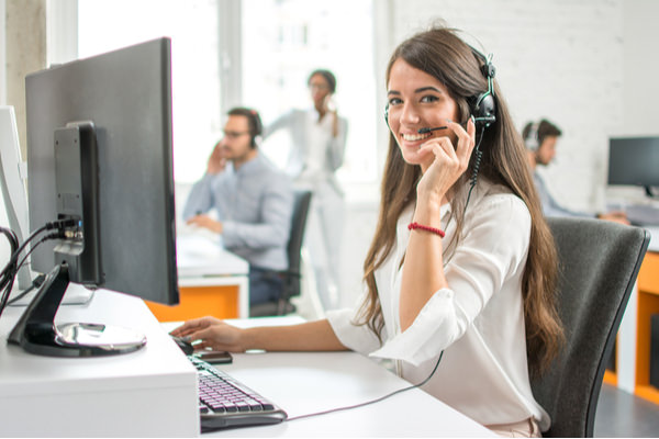 A woman is at her computer, she is smiling at the camera and has a headset on.