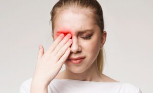 A blonde woman rubs her right eye, and the area around her eye is glowing red, indicating pain